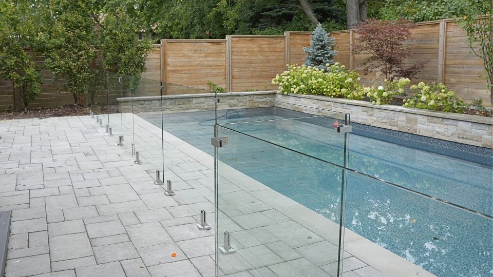 Frameless glass pool fence with stainless steel spigots installed on a stone patio around a backyard inground pool