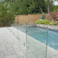 Glass pool railing system with stainless steel base spigots enclosing a backyard pool, surrounded by a stone deck and landscaped garden